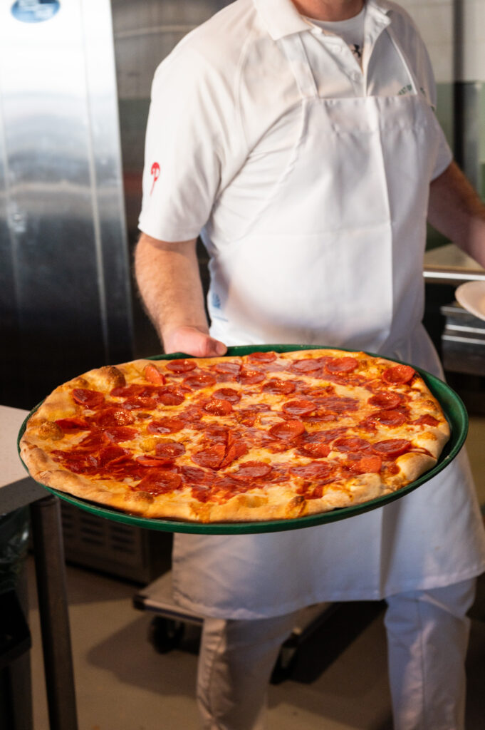 A Manco & Manco pieman carrying a fresh pepperoni pie out to the counter